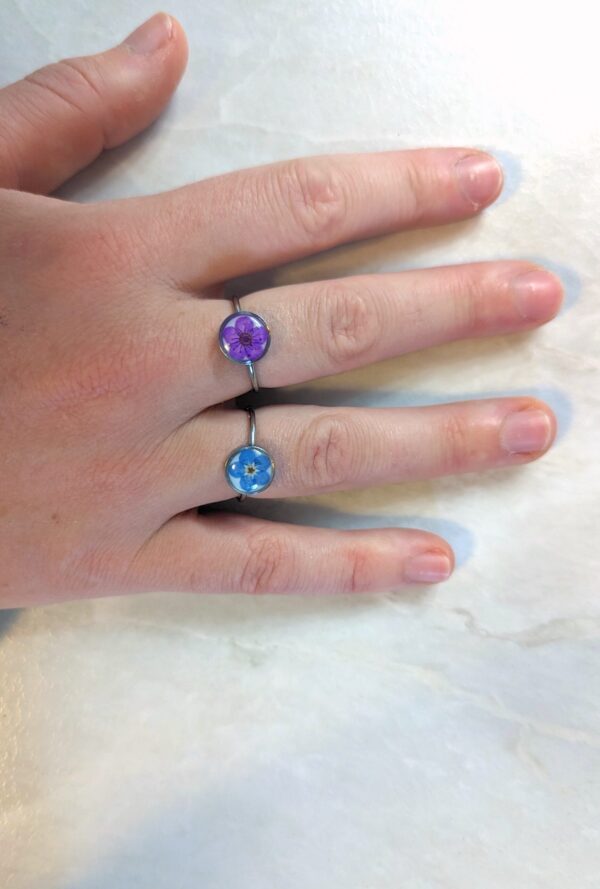 a hand with two real-flower rings on - both silver with circle frames and a purple and blue flower inside