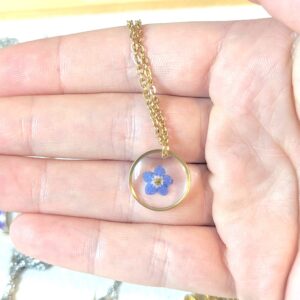 a hand holding a gold dainty necklace with a circle pendant with a little forget-me-not flower pressed inside a clear window