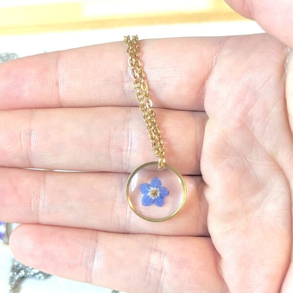a hand holding a gold dainty necklace with a circle pendant with a little forget-me-not flower pressed inside a clear window