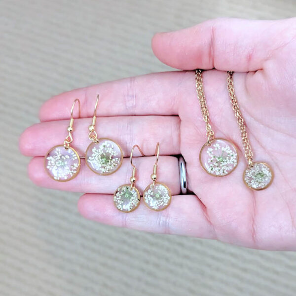 a hand holding earrings and necklaces with real pressed white wildflowers inside