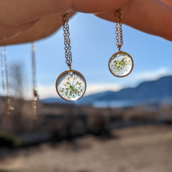 a sunny autumn day overlooking a lake and mountains, in the foreground a hand is dangling 2 real pressed flower necklaces
