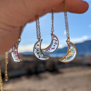 a sunny autumn day overlooking a lake and mountains, in the foreground a hand is dangling 4 wildflower moon necklaces